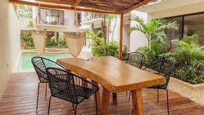 Ein Holztisch mit vier Stühlen unter einem Reetdach, umgeben von üppiger Vegetation und einem Pool.
