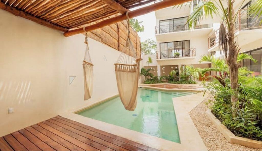 Un espace piscine doté d’une terrasse en bois, de fauteuils suspendus et d’un bâtiment avec des balcons et des plantes.