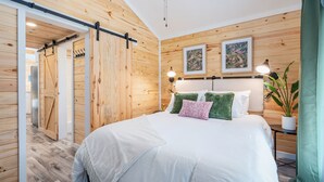 Ein Schlafzimmer mit einer Wand aus Holzlatten, einem Bett mit weißer Bettwäsche und grünen und pinkfarbenen Kissen, einem Nachttisch mit Lampe und einer Topfpflanze.