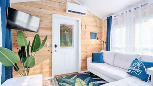 Ein modernes Wohnzimmer mit einem weißen Sofa, einem flachbildfernseher an der Wand, einer Holzbalkendecke und einem großen Fenster mit durchsichtigen Vorhängen.