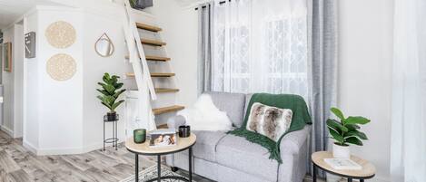A modern living room with a staircase, a grey sofa, a small round table, and two potted plants.
