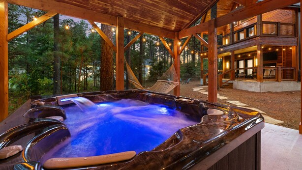 A hot tub with a wooden deck and a hammock, surrounded by a forest and a building with a balcony. - Mojave in the Pines - New Luxury Cabin in Hochatown Sleeps 4 (Broken Bow)