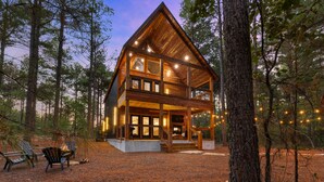 A two-story log cabin with a balcony, surrounded by trees and a gravel driveway. - Mojave in the Pines - New Luxury Cabin in Hochatown Sleeps 4 (Broken Bow)