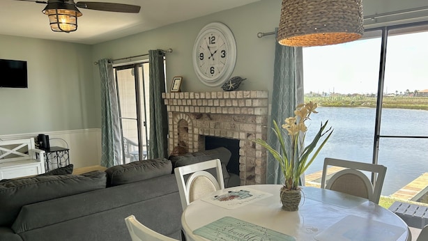 A living room with a fireplace, a dining table with chairs, a sofa, and a large window overlooking a body of water.