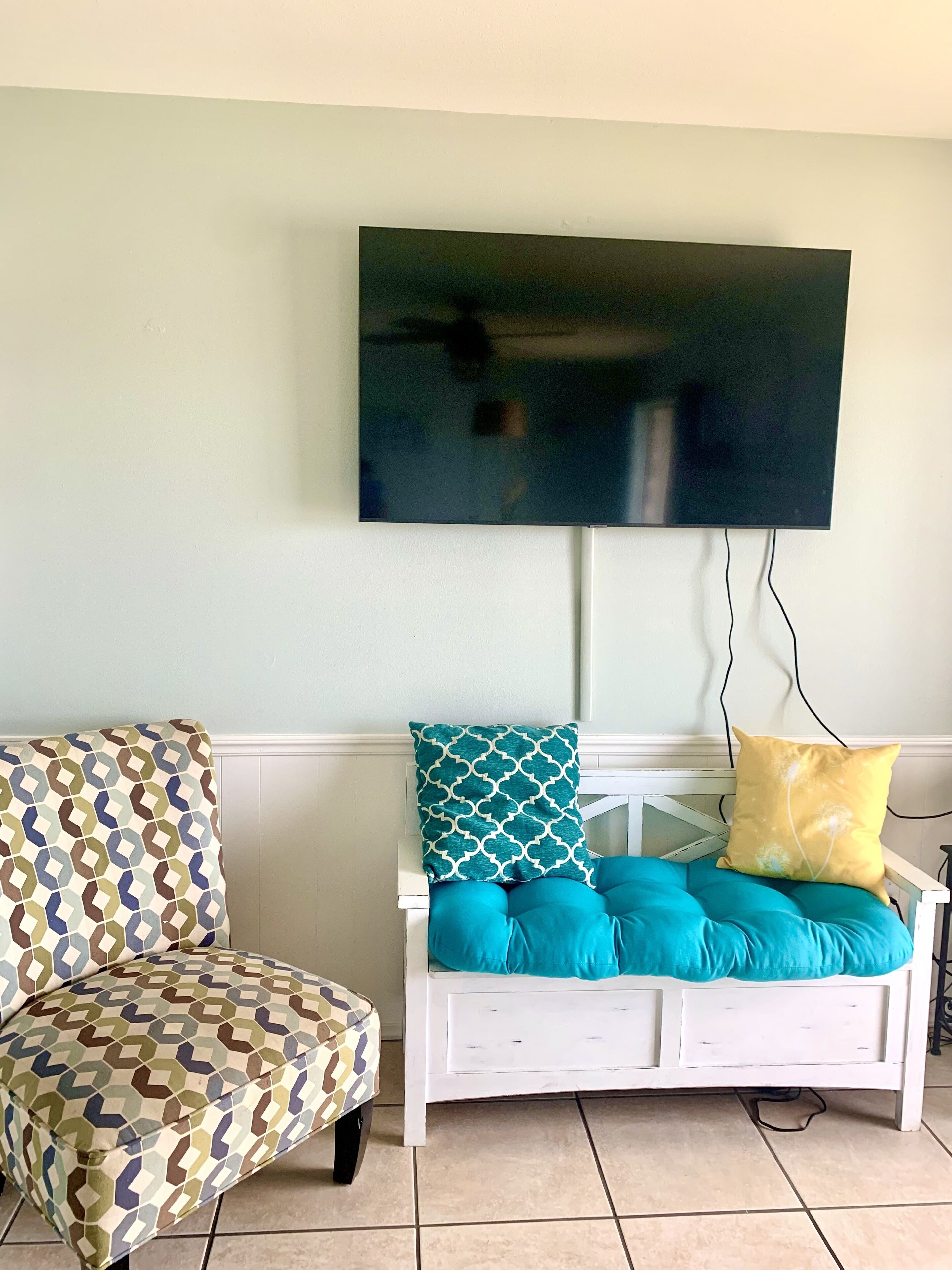 A living room with a white sofa, patterned armchair, and a flat-screen TV mounted on the wall.