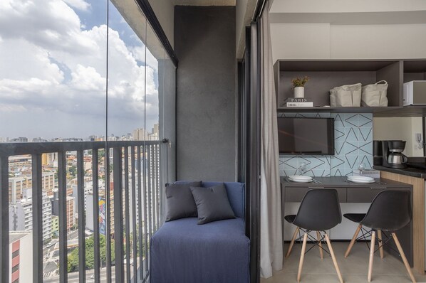 A balcony with a city view, a sofa with cushions, a dining area with chairs, and a kitchen area with a coffee machine.