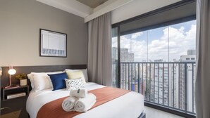 Una habitación de hotel moderna con una cama grande, una mesita de noche con lámpara, una obra de arte enmarcada y vistas a la ciudad a través de amplios ventanales.