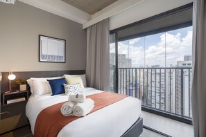 Una habitación de hotel moderna con una cama grande, una mesita de noche con lámpara, una obra de arte enmarcada y vistas a la ciudad a través de amplios ventanales.