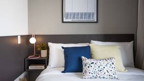 Una cama bien hecha con ropa de cama blanca, un almohadón azul, un almohadón amarillo y un almohadón con estampado multicolores. Encima de la cama hay un cuadro con texto, una mesita de noche con una lámpara rosa y una planta en maceta.