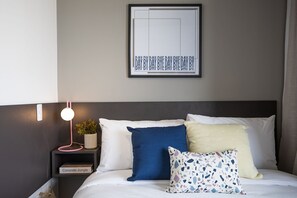 Ein ordentlich gemachtes Bett mit weißer Bettwäsche, einem blauen Kissen, einem gelben Kissen und einem mehrfarbigen Kissen mit Muster. Darüber hängt ein gerahmtes Bild mit Text, neben dem Bett steht ein kleiner Nachttisch mit einer pinkfarbenen Lampe und eine Topfpflanze.