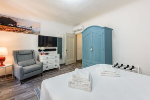 Habitación de hotel con una cama, televisión, armario azul, sillón gris y un cuadro en la pared.