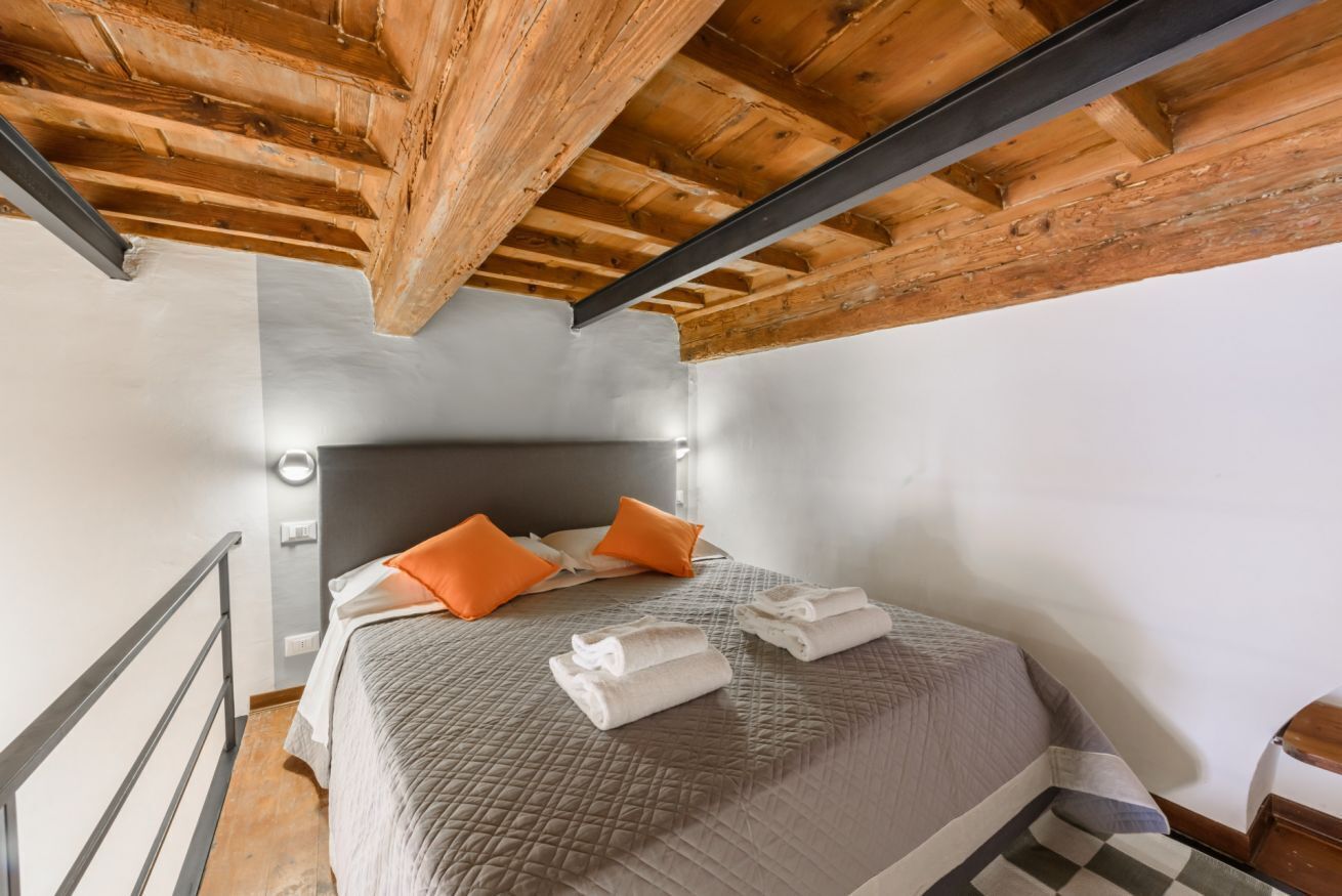 Una camera da letto con soffitto in legno, un letto con copriletto grigio e cuscini arancioni, e un poggiatesta in metallo.