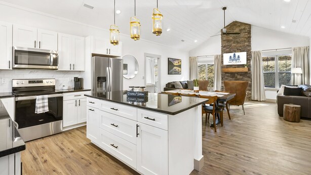 Una cocina moderna con armarios blancos, electrodomésticos de acero inoxidable y una isla central. Hay un comedor con una mesa y sillas de madera, y una sala de estar con un sofá y una chimenea de piedra.