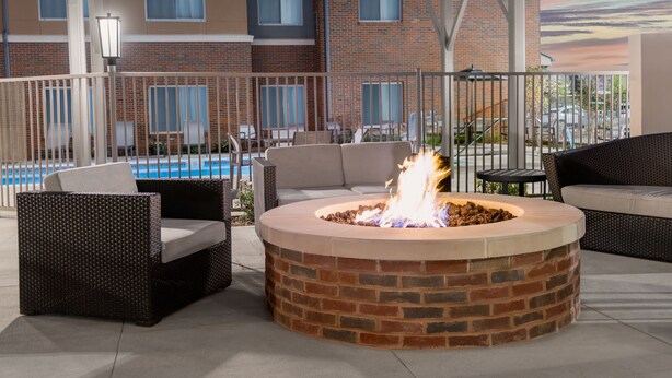 Una chimenea circular con base de ladrillo rodeada de mobiliario de mimbre, incluyendo sofás y sillones, en un patio al aire libre con una piscina al fondo.
