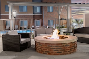 Una chimenea circular con base de ladrillo rodeada de mobiliario de mimbre, incluyendo sofás y sillones, en un patio al aire libre con una piscina al fondo.