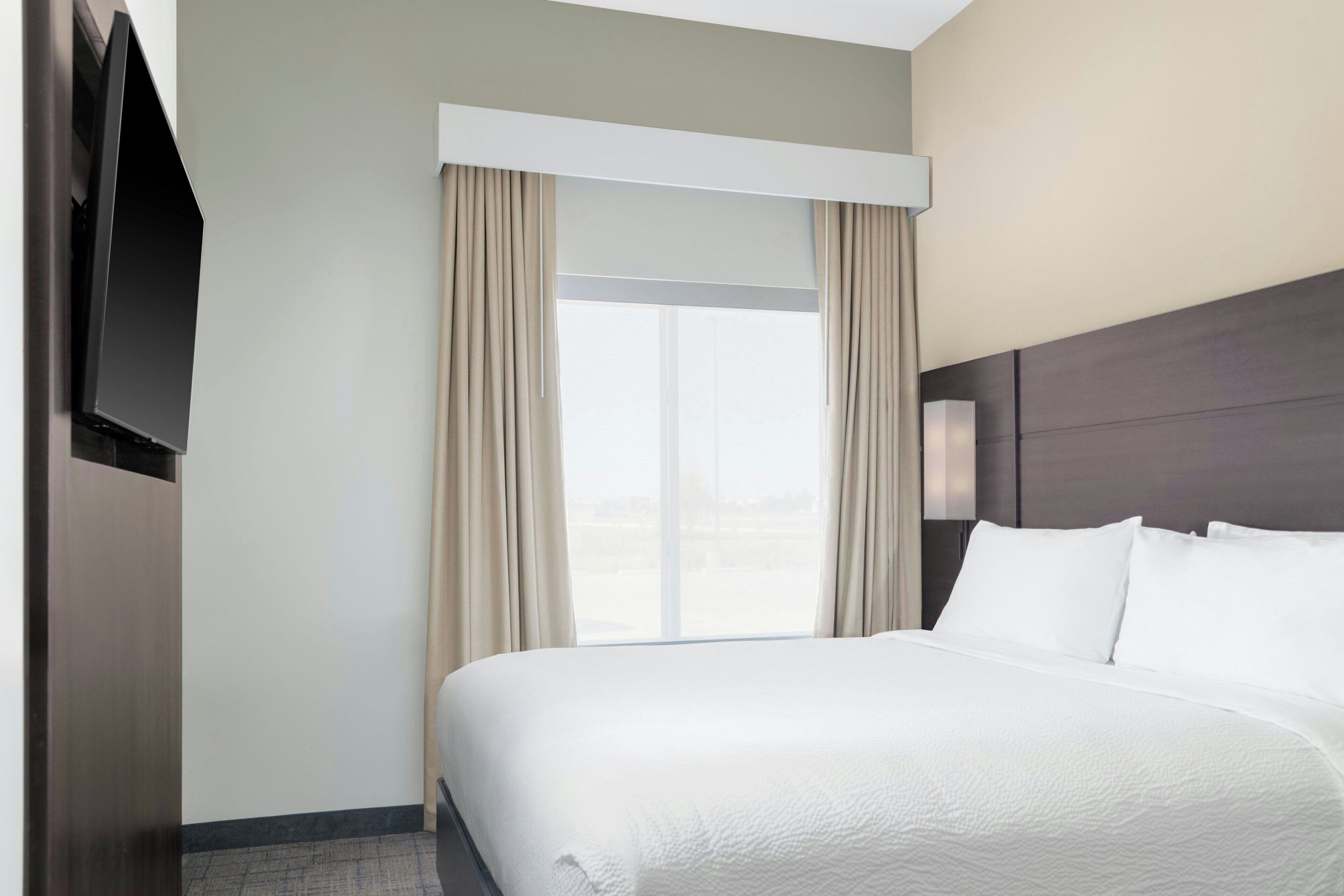 A hotel room with a large bed, a flat-screen TV mounted on the wall, and a window with curtains.