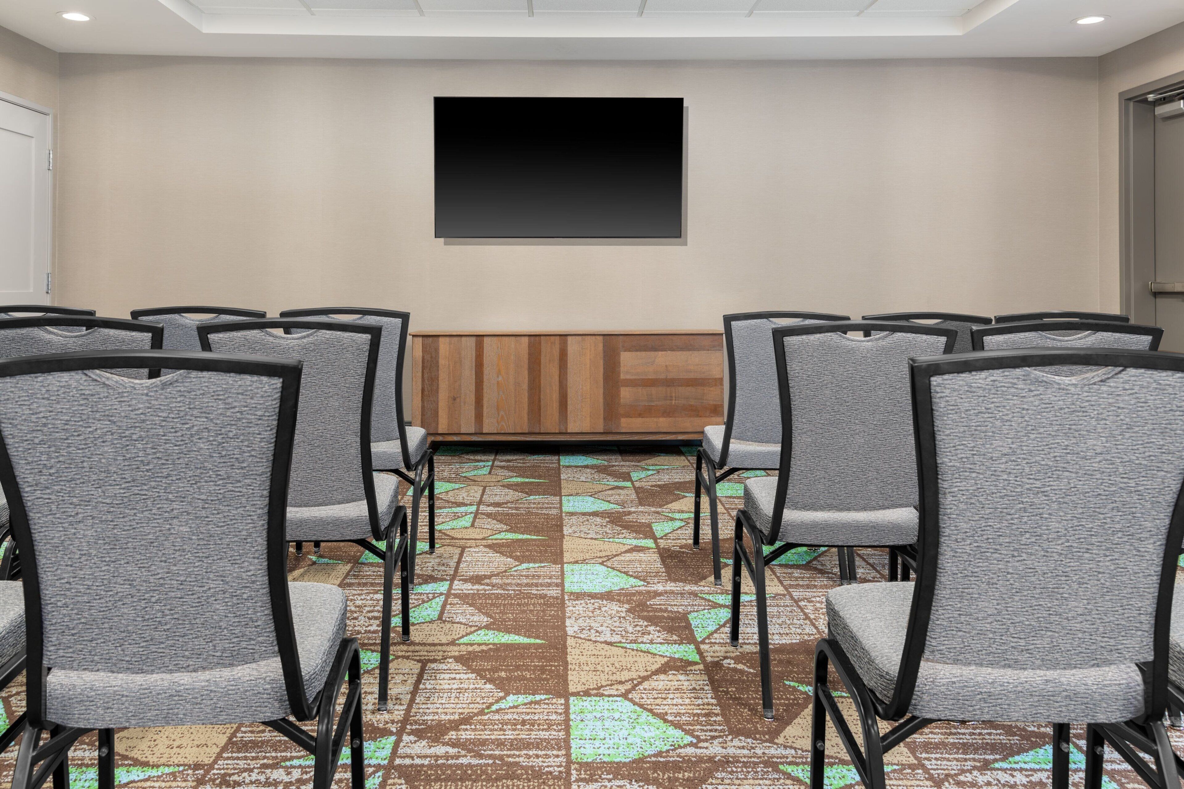 Uma sala de conferências com fileiras de cadeiras cinzas, um painel de parede em madeira e um tapete estampado.
