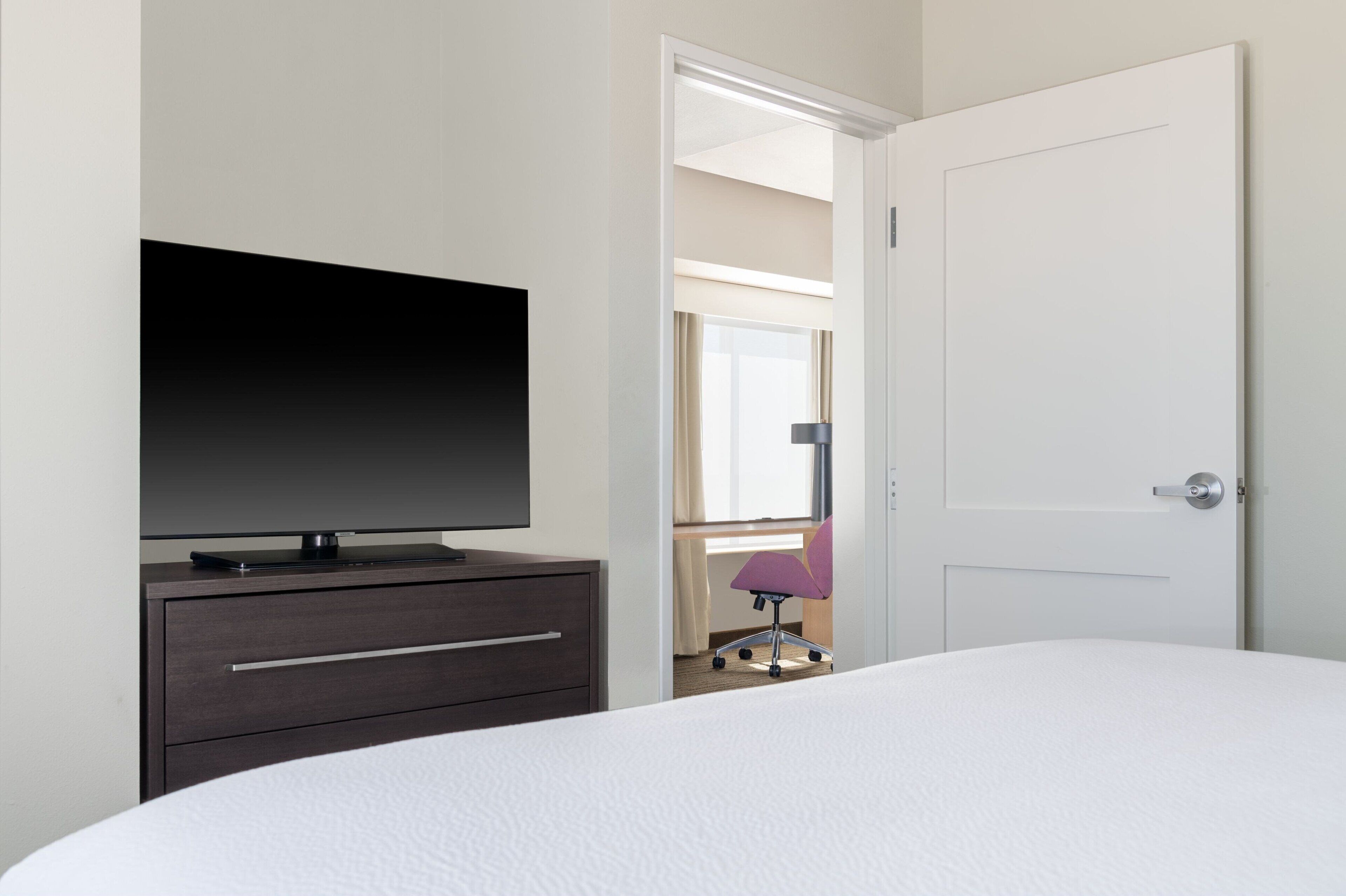 A hotel room with a bed, a TV on a dresser, a door, and a chair in the background.