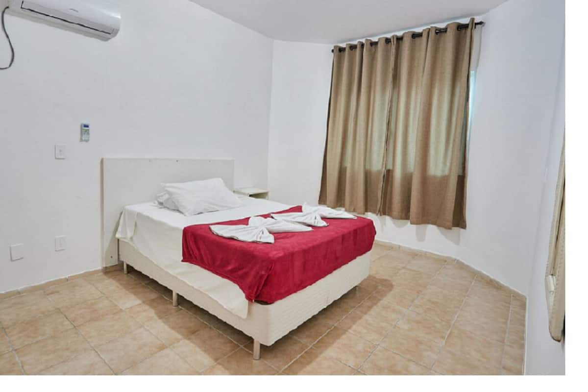 A hotel room with a bed, white walls, a window with curtains, and a tiled floor.