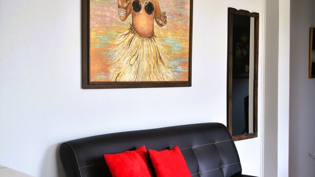 A black leather sofa with two red cushions, a framed painting of a woman in a tropical setting, and a mirror on the wall. - Papaya Dreams (San Juan)