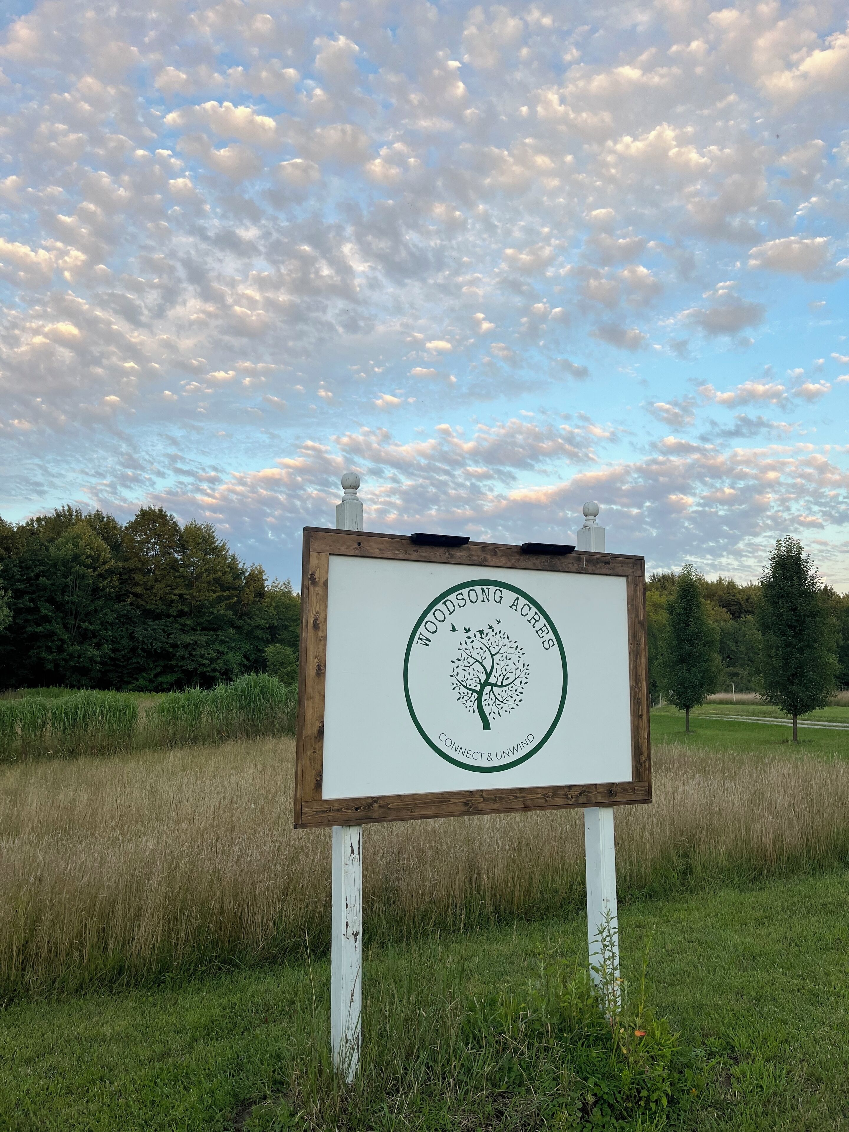 Un letrero con un logo de árbol y el texto 'Woodsong Acres Connect & Unwind' en un campo cubierto de hierba.