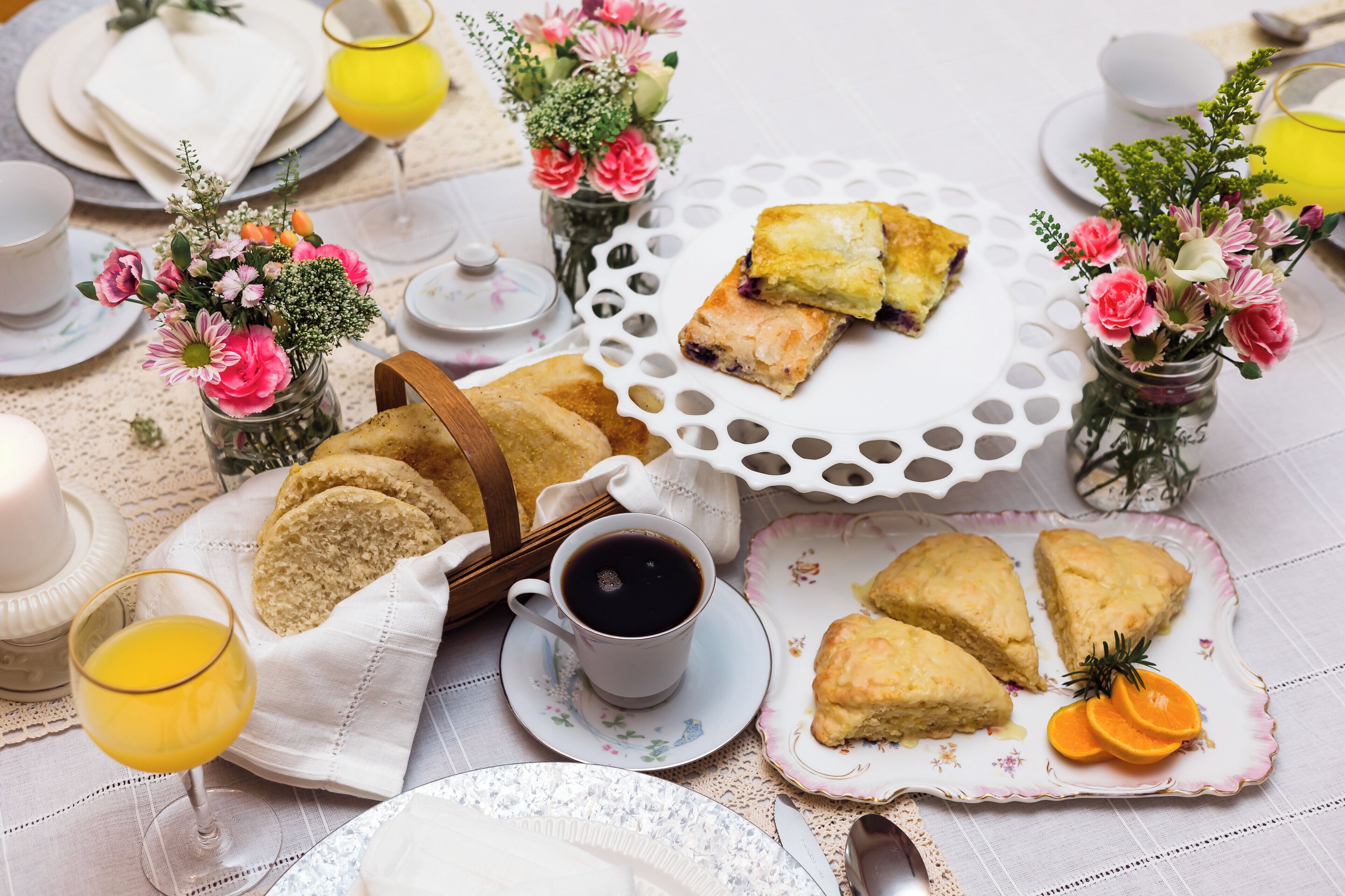 Un desayuno con bollos, café y jugo sobre una mesa con manteles de encaje y centros de mesa florales.