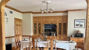 A dining area with a wooden table and chairs, a television above the table, and wooden cabinets in the background. - The Inn at Woodsong Acres (Kingsville)