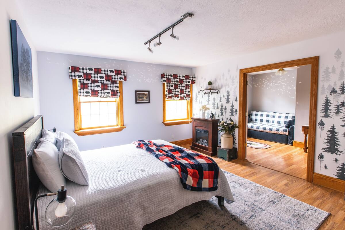 A bedroom with a bed, two windows with patterned curtains, a fireplace, and a view into another room.