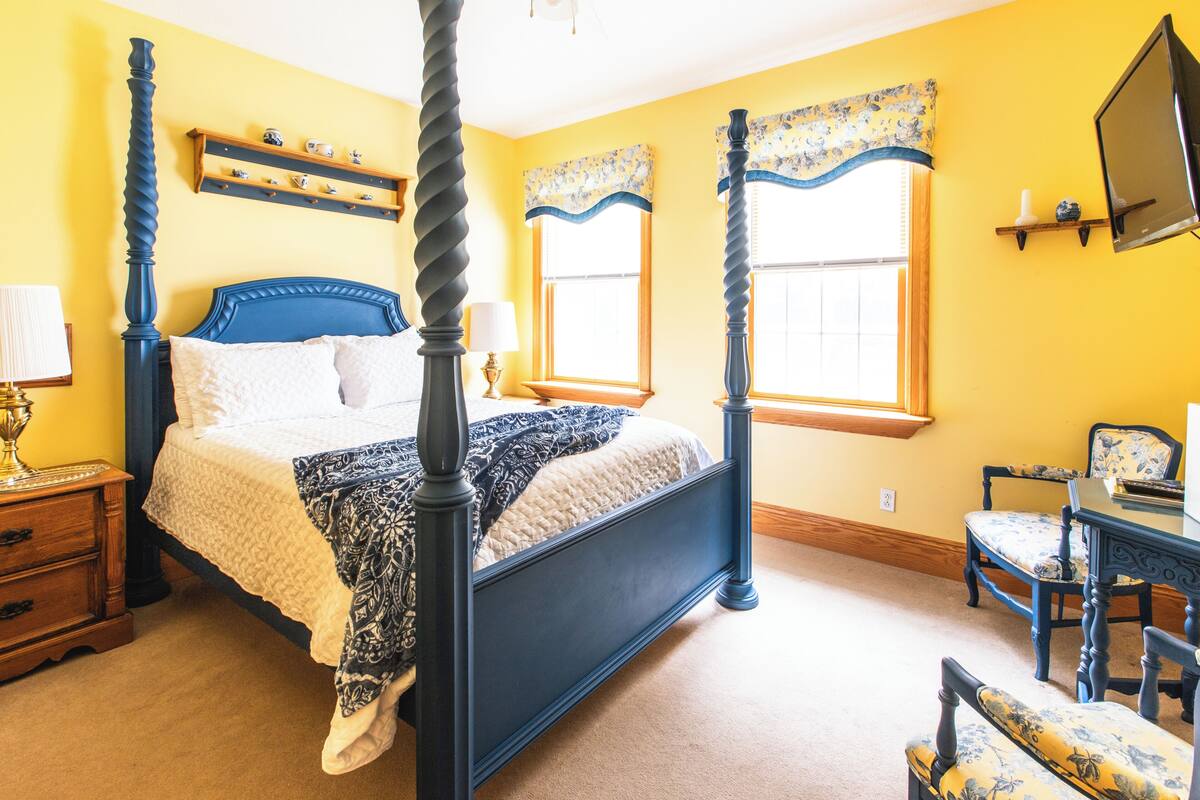 A four-poster bed with a dark blue frame, a bedside table with a lamp, a chair with floral upholstery, and a television mounted on the wall.
