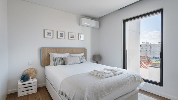 Ein modernes Schlafzimmer mit einem großen Bett, einem Nachttisch, einer Lampe und Blick auf die Stadt durch ein großes Fenster.