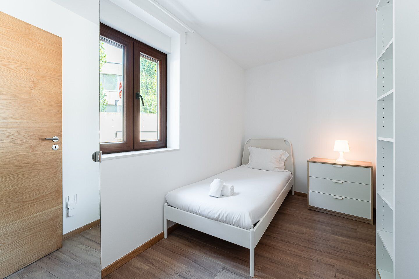 A small, neatly arranged bedroom with a single bed, a bedside table with a lamp, and a wardrobe.
