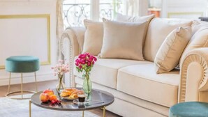 A cozy living room with a beige sofa, a round coffee table with fruits and pastries, and a vase with flowers.