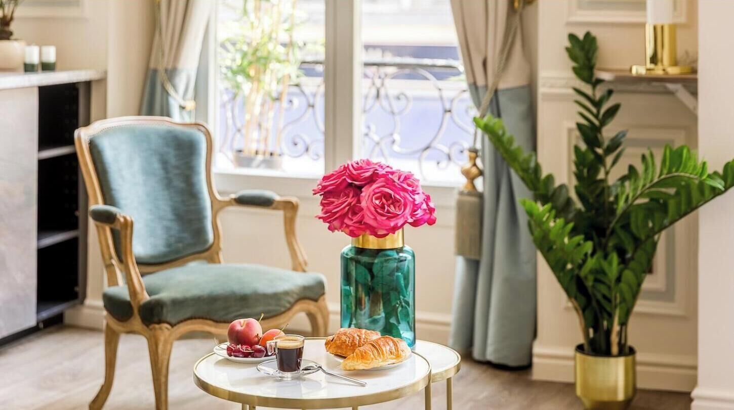 A dining table with a vase of flowers, a plate of fruit, a croissant, and a cup of coffee.