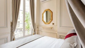 A hotel room with a bed, a large window with curtains, a gold-framed mirror, and a view of greenery outside.