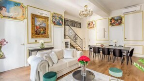 A spacious living room with a sofa, dining table, and chairs, a staircase, and decorative paintings on the walls.