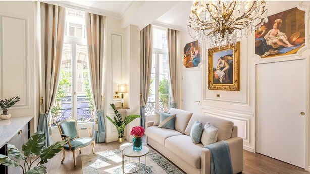 A well-lit living room with a beige sofa, a green armchair, and a chandelier. There are paintings on the walls and a window with curtains.