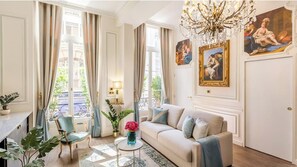Un soggiorno luminoso con un divano color beige, una poltrona verde e un lampadario. Alle pareti si notano dei dipinti e una finestra con tende.