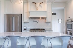 Cozinha moderna com armários brancos, uma ilha central com banquetas, geladeira de aço inoxidável, fogão e um crânio de vaca fixado na parede.