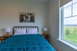 A bedroom with a bed, two bedside lamps, a window with a view, and a painting on the wall.