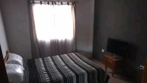 Un dormitorio con una cama, un televisor sobre un soporte de madera y una ventana con cortinas.