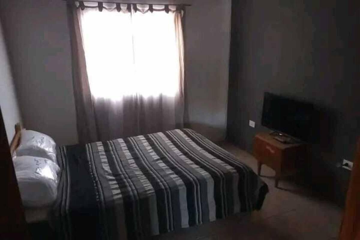 A bedroom with a bed, a television on a wooden stand, and a window with curtains.