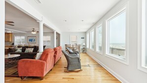 A spacious living room with a wooden floor, a red sofa, a grey rocking chair, and a view of the ocean through large windows.