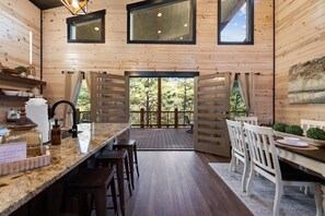 A wooden cabin with a kitchen area featuring a marble countertop, bar stools, and a dining table with chairs. There are large windows and a view of the surrounding forest. - Pets, Big Deck, Hot Tub, Fire Pit, Mountainside Escape (Broken Bow)