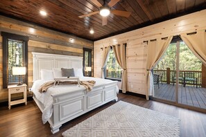 A large bed with a white headboard, a wooden ceiling fan, and a view of a wooden deck through a window. - Pets, Big Deck, Hot Tub, Fire Pit, Mountainside Escape (Broken Bow)