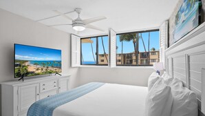 Una camera da letto con un letto grande, un ventilatore a soffitto, una TV a parete e una finestra che si affaccia su palme e edifici.