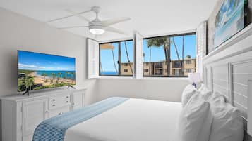 A bedroom with a large bed, a ceiling fan, a TV on the wall, and a window with a view of palm trees and buildings.