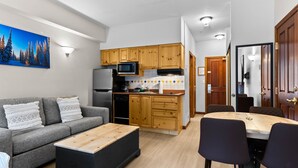 A modern kitchen with wooden cabinets, a microwave, and a refrigerator. There is a dining table with chairs and a gray sofa with decorative pillows.