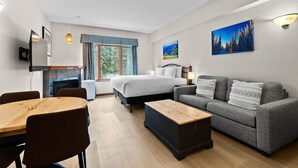 A modern hotel room with a bed, sofa, dining table, and a small kitchen area.