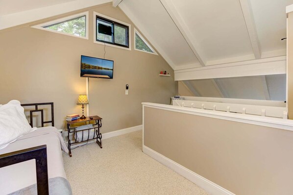 A bedroom with a bed, a desk with a lamp, a TV mounted on the wall, and a window with a view of trees.