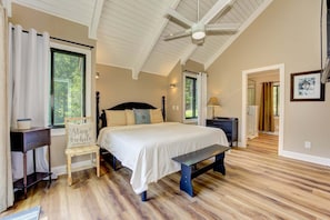 A bedroom with a large bed, a desk, a chair, and a bench. There are windows and a ceiling fan.